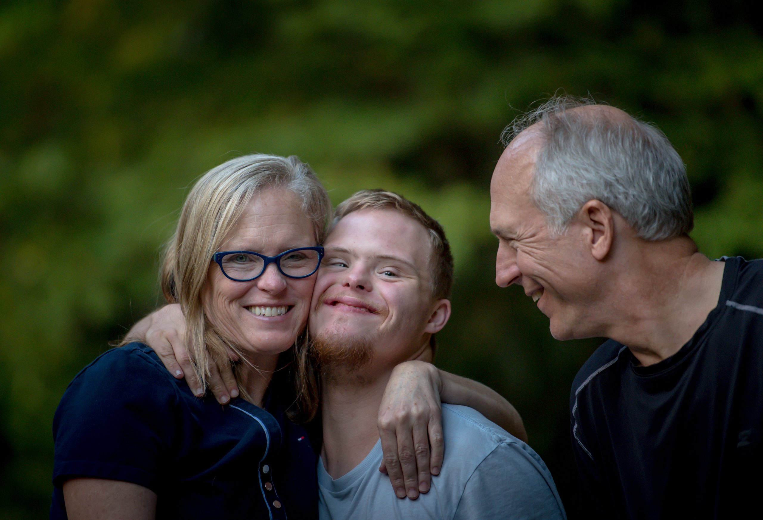 picture of a family outdoors with a mom with blonde hair, blue-rimmed glasses and navy short sleeve shirt is smiling and has her arm around her son, also smiling, with light hair wearing a light tee-shirt and has his arm around his mom while the dad with gray hair, also wearing a navy blue shirt, looks at them smiling